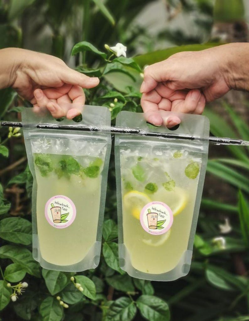 May include: Two clear plastic pouches with a straw hanging out of the top. The pouches contain a light yellow liquid with green mint leaves and lemon slices. The pouches have a pink sticker with the text "Winkwink Tea" and a cartoon teacup with a face.