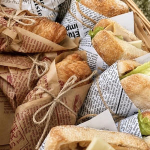 Könnte beinhalten: Eine Ansammlung von Sandwiches und Croissants, eingewickelt in weißes und braunes Papier, das mit schwarzem französischem Zeitungstext bedruckt ist. Jeder Artikel ist mit dünner Jutekordel gebunden. Die Sandwiches enthalten grünen Salat und Käse.