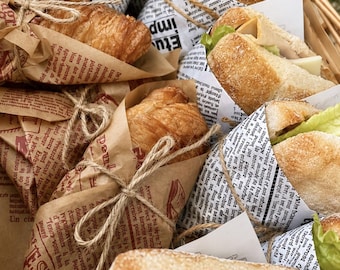 Puede incluir: Una colección de sándwiches y croissants envueltos en papel blanco y marrón impreso con texto de periódico francés en negro. Cada artículo está atado con cuerda de yute fina. Los sándwiches contienen lechuga verde y lonchas de queso pálido.