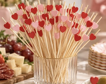 May include: A collection of wooden food picks featuring small red and light pink heart-shaped toppers. The picks are arranged upright in a clear, vertically ribbed glass container, creating a dense cluster of decorative heart accents.