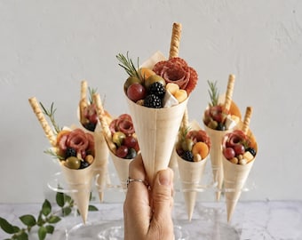 May include: A collection of charcuterie cones served in light wood veneer holders. Each cone contains a rolled slice of salami, a wedge of cheese, a blackberry, a green olive, a red grape, a sprig of rosemary, and a thin, textured breadstick.
