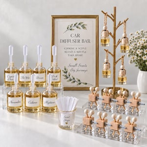 May include: A car diffuser bar display featuring glass bottles with droppers, labeled with scent names like Crystal Clear and November. A gold tree stand holds hanging diffusers, while a sign reads Car Diffuser Bar, Choose a Scent, Blend a Bottle, Take Home.