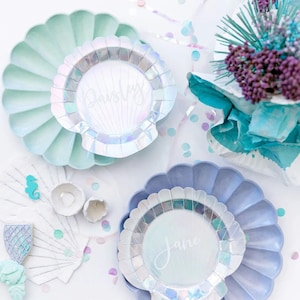 May include: Two iridescent white seashell-shaped paper plates with scalloped edges. The plates are on a white background with confetti and a blue and green floral arrangement. The top plate has the name "Paisley" written on it. The bottom plate has the name "Jane" written on it.
