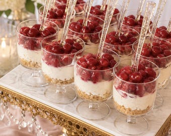 May include: A collection of clear plastic dessert cups on a gold pedestal stand. Each cup contains a bottom layer of crushed graham cracker, a middle layer of white cream, and a top layer of red cherries, finished with a clear glittery spoon.
