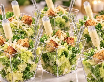 May include: Several clear square plastic cups filled with chopped romaine lettuce, shredded parmesan cheese, and toasted croutons. Each cup includes a small clear plastic pipette filled with creamy dressing and a clear plastic tasting fork.