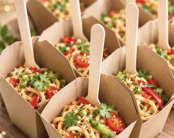 May include: Multiple individual servings of noodles in small, square, light brown paper containers. Each container holds a portion of noodles topped with sliced red cherry tomatoes, green herbs, and sesame seeds, with a wooden spoon inserted.