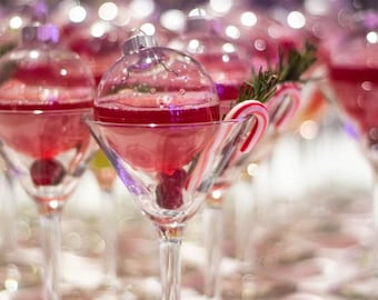 Könnte beinhalten: Eine klare, kugelförmige Glaskugel, gefüllt mit einer leuchtend roten Flüssigkeit, die in einem Martiniglas mit Stiel ruht. Eine kleine Zuckerstange und ein Zweig grüner Rosmarin stecken im Glas, eine dunkle Kirsche ist am Boden sichtbar.