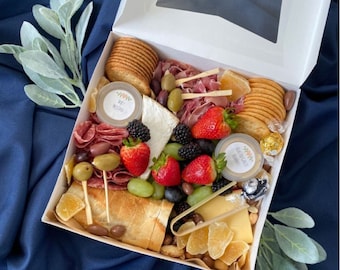 May include: A white box filled with a gourmet charcuterie board. The board includes crackers, cheese, fruit, meat, olives, and nuts. The box has a clear plastic window on the top.
