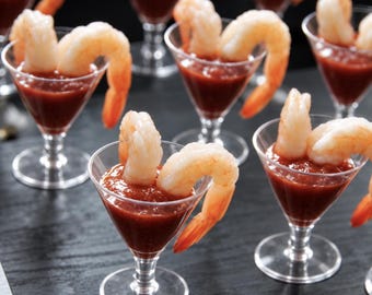 May include: Multiple clear plastic martini-style glasses filled with a thick red cocktail sauce and garnished with a single large, cooked, pink and white shrimp draped over the rim of each individual serving vessel.