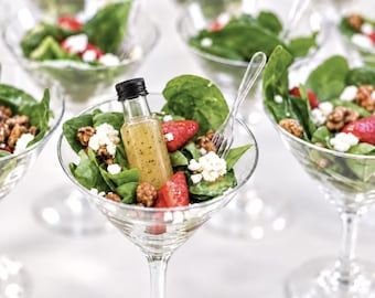 May include: A clear martini glass filled with a salad of fresh green spinach leaves, sliced red strawberries, and white crumbled cheese. A small glass bottle with a black cap containing salad dressing sits inside the glass.