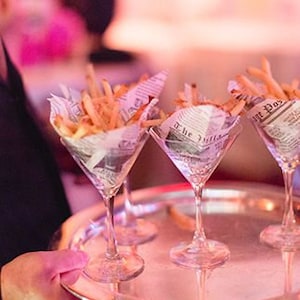 May include: A silver tray holds four clear glass martini glasses filled with french fries wrapped in newspaper. The glasses are on a white surface.