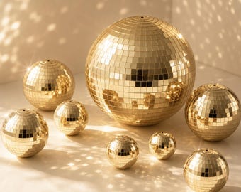 May include: A collection of eight spherical disco balls in varying sizes, each covered in small, reflective gold-colored square mirror tiles. The spheres are arranged in a group, creating a uniform metallic appearance with light reflecting off the surfaces.
