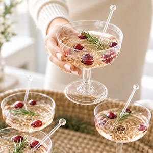 May include: Clear glass coupe cocktail glasses filled with a pale golden liquid, each garnished with fresh red cranberries and a sprig of green rosemary. A transparent stirring rod with a spherical top is placed inside each glass.