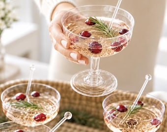 May include: Clear glass coupe cocktail glasses filled with a pale golden liquid, each garnished with fresh red cranberries and a sprig of green rosemary. A transparent stirring rod with a spherical top is placed inside each glass.