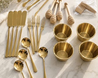May include: A collection of gold-toned metal serving utensils including four butter knives, four forks, four spoons, two tongs, and four small bowls, accompanied by four wooden honey dippers and a small plastic bag of wooden toothpicks.