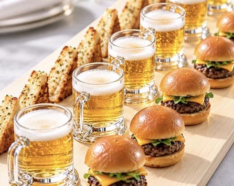 May include: A wooden serving board features a row of small glass beer mugs filled with golden liquid and white foam, accompanied by a line of toasted bread triangles and several small cheeseburger sliders with green lettuce.
