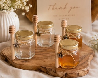 May include: Four hexagonal glass jars with gold lids, each paired with a wooden honey dipper. Two jars are filled with amber honey, while two are empty. Each jar features a small metal bee charm tied with twine around the neck of the glass.