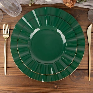 May include: A set of three emerald green plates with ruffled edges and gold trim. A gold fork and knife are placed on a wooden surface, alongside a glass and decorative elements, suggesting a formal dining setting.
