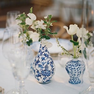 Op de afbeelding: Twee kleine, wit en blauw porseleinen vazen met bloemmotieven bevatten witte bloemen. De vazen staan op een wit tafelkleed, met helder glaswerk op de voorgrond. De vazen hebben een klassieke, elegante uitstraling.