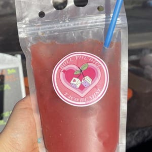 May include: A clear plastic pouch with a pink and white sticker that says "Roll the Dice Taco Remix" and a red liquid inside. A blue straw is sticking out of the top of the pouch.