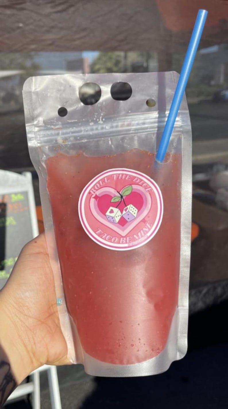 May include: A clear plastic pouch with a pink and white sticker that says "Roll the Dice Taco Remix" and a red liquid inside. A blue straw is sticking out of the top of the pouch.