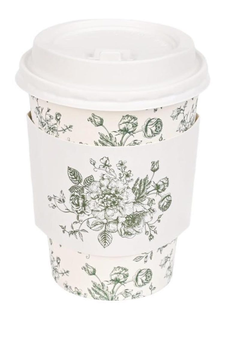May include: A white disposable coffee cup with a matching lid. The cup has a floral design in green and white, with a matching white sleeve. The floral pattern includes roses and other flowers.
