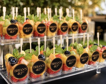 May include: Clear square plastic cups filled with fresh fruit salad, including strawberries, blackberries, and melon, garnished with mint leaves and wooden skewers. Each cup features a round black sticker with gold text reading BRIAN'S 40TH.