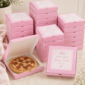 Op de afbeelding: Een stapel vierkante roze kartonnen pizzadozen met witte etiketten. Elk etiket bevat de tekst 'Thank you for celebrating', 'Skinny Bitch PIZZA' en een klein hartje. Eén open doos bevat een ronde pepperoni-pizza in de verpakking.