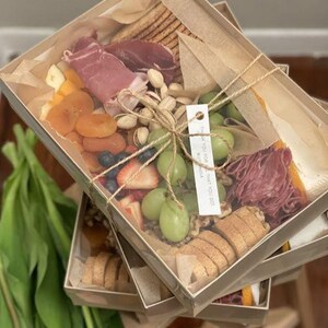 Puede incluir: Varias cajas con tapa transparente llenas de charcutería. Las cajas contienen galletas saladas, embutidos, quesos, albaricoques secos, uvas, fresas, arándanos y pistachos. Cada caja está atada con hilo y tiene una pequeña etiqueta.