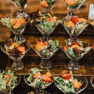 May include: A tiered display of small glass bowls filled with salad. The salad is made with fresh greens, orange slices, strawberries, and a white cheese crumbles.