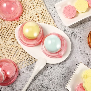 Puede incluir: Dos recipientes de plástico transparente con tapas rosas, cada uno con un macaron de color pastel. Los recipientes están en un plato blanco con una cuchara blanca. Los macarons son amarillos y azules.