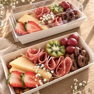 Op de afbeelding: Twee rechthoekige witte papieren voedselbakjes gevuld met een assortiment van gesneden salami, gele kaaspartjes, verse aardbeienhelften, kiwischijfjes, rode druiven, met chocolade bedekte pretzels en amandelen, gegarneerd met kleine witte bloemen.