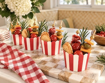 May include: Four individual snack cups featuring red and white vertical stripes, each filled with a skewer of cheddar cheese, a green olive, a cherry tomato, and folded salami, accompanied by round crackers and a sprig of fresh rosemary.
