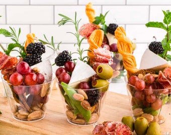 Puede incluir: Tres vasos de plástico transparente llenos de una variedad de aperitivos, que incluyen uvas, queso, salami, aceitunas, nueces y galletas. Los vasos están dispuestos sobre una superficie de madera.
