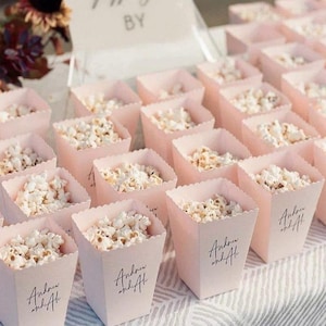Op de afbeelding: Rijen lichtroze popcornbakjes gevuld met popcorn. Elk bakje heeft een geschulpte rand en de tekst "Andrew and Ali" in zwart schrift. De bakjes staan op een witte ondergrond.