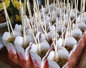 Puede incluir: Una bandeja de color cobre con numerosas cajas individuales de comida para llevar china, cada una con fideos y verduras, con palillos. Las cajas son blancas con caracteres chinos rojos. Los palillos son de madera clara.