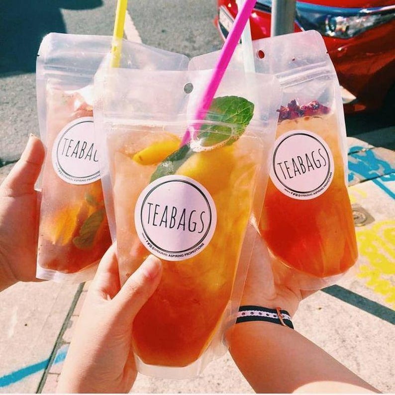 May include: Three clear plastic pouches filled with a refreshing drink. The pouches have a white circular label with the text "TEABAGS" printed in black. The drink is a light orange colour and contains fruit slices and mint leaves.