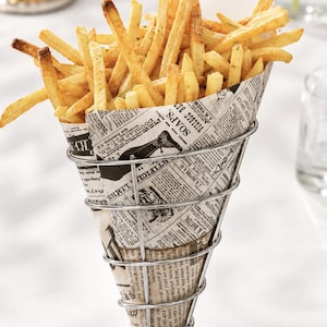 May include: A serving of golden-brown french fries held in a cone-shaped paper liner with vintage newspaper print, placed inside a silver-toned wire metal stand with a circular base. The paper features black text and illustrations of antique items.