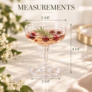 May include: A clear glass coupe cocktail glass filled with a sparkling pink beverage, cranberries, and a rosemary sprig. The glass features a stem and circular base, with dimensions labeled as 3 3/8 inches wide, 4 1/4 inches tall, and a 2 1/4 inch base.
