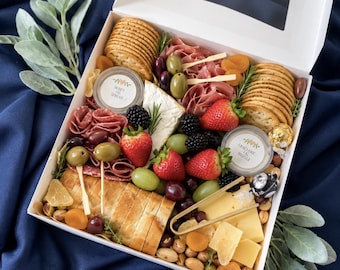 May include: A white gift box containing a charcuterie arrangement with round crackers, sliced baguette, brie cheese, cheddar cheese, salami, prosciutto, strawberries, blackberries, green and purple grapes, olives, dried apricots, nuts, and two small containers.