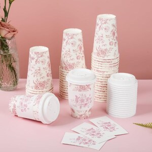 May include: A selection of disposable coffee cups and lids, patterned with pink and white floral designs. The cups are in different sizes and arrangements, some stacked, others lying down. Small cards with the same floral design are also visible.