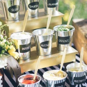 Puede incluir: Un soporte de varios niveles con pequeños cubos de metal llenos de diferentes ingredientes para hot dogs. Los cubos están etiquetados con letreros de pizarra: "Relish", "Tomates", "Jalapeños", "Cebollas", "Mostaza", "Ketchup", "Queso" y "Mostaza".