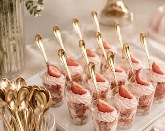 May include: A collection of small clear plastic cups filled with layers of cake and pink cream, each topped with a fresh strawberry slice and a gold-toned metal spoon. A glass container holds a cluster of additional gold-toned metal spoons.