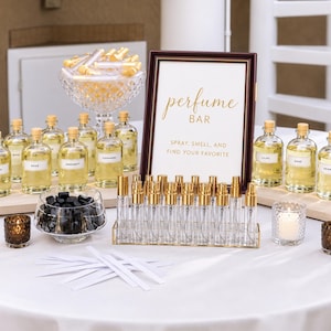 May include: A perfume bar display featuring a framed sign that reads perfume BAR SPRAY, SMELL, AND FIND YOUR FAVORITE. The setup includes multiple glass bottles filled with yellow liquid, a crystal bowl of small vials, and a gold rack of atomizers.
