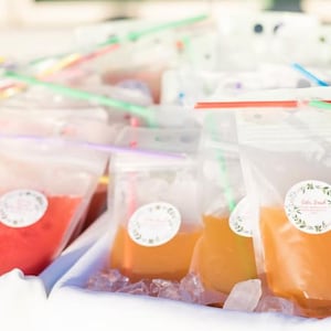 May include: A close-up of a table filled with clear plastic bags filled with colourful liquids. The bags have labels with the text "Little Sips" on them. The bags are arranged in rows and are surrounded by ice.