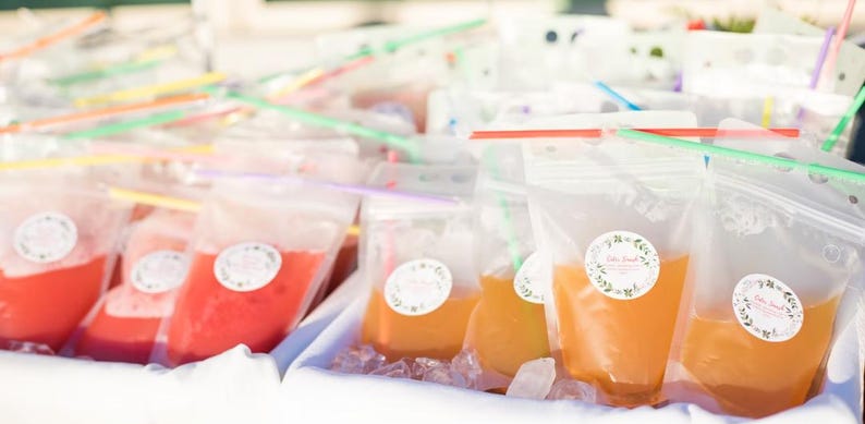 May include: A close-up of a table filled with clear plastic bags filled with colourful liquids. The bags have labels with the text "Little Sips" on them. The bags are arranged in rows and are surrounded by ice.