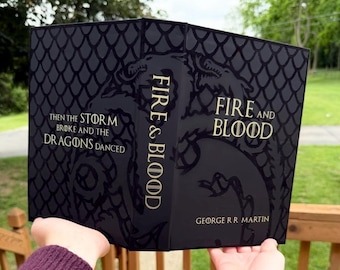Puede incluir: Un libro de tapa dura negro titulado Fire and Blood de George R.R. Martin. La portada presenta un patrón brillante de escamas de dragón, una gran silueta de dragón y texto dorado que dice: Then the storm broke and the dragons danced.