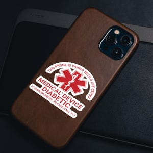 May include: A brown leather phone case with a sticker that reads "THIS PHONE IS PAIRED WITH A LIFE-SAVING MEDICAL DEVICE DIABETIC IF UNRESPONSIVE, PLEASE CALL 911." The phone has a triple-lens camera.