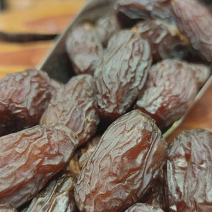 May include: A metal scoop filled with a heap of dried brown dates. The dates have a wrinkled, textured skin with a deep reddish-brown colour and an elongated, oval shape. The fruit is piled high in the metallic container, showing natural variations.