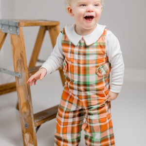 May include: A toddler wearing a white long-sleeved shirt and orange, green, and white plaid overalls. The overalls have a white collar and decorative buttons. A wooden ladder is in the background.